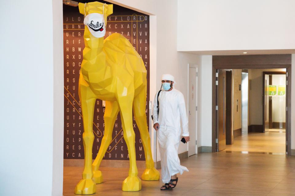 An Emirati wearing a face mask due to the coronavirus pandemic walks past a camel statue decorated with a face mask at the Rove City Centre Hotel in Dubai, United Arab Emirates, Monday, July 6, 2020. Dubai reopened for tourists Tuesday amid the coronavirus pandemic, hoping to reinvigorate a vital industry for this city-state before its crucial winter tourist season. (AP Photo/Jon Gambrell)