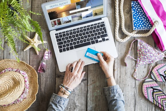 Woman holding credit card looking at a travel agency on a laptop, with a hat, swimsuit, beach tote, and flip-flops surrounding the computer