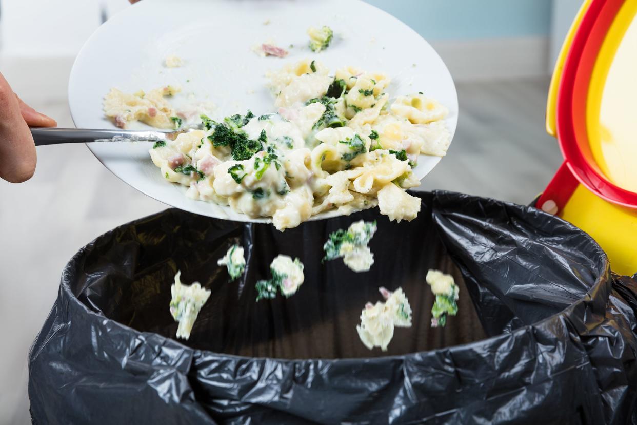 Closeup of a person's hands scraping leftover pasta off of a white plate into the trash bin, with grey floor and blue walls in the background