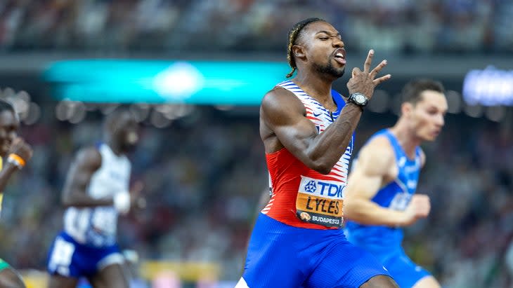 A strinter in red white and blue singlet holds up three fingers for the victories