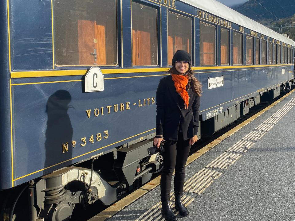 The author wearing black stands on a platform in front of a navy blue train with gold accents. Mountains are in the background