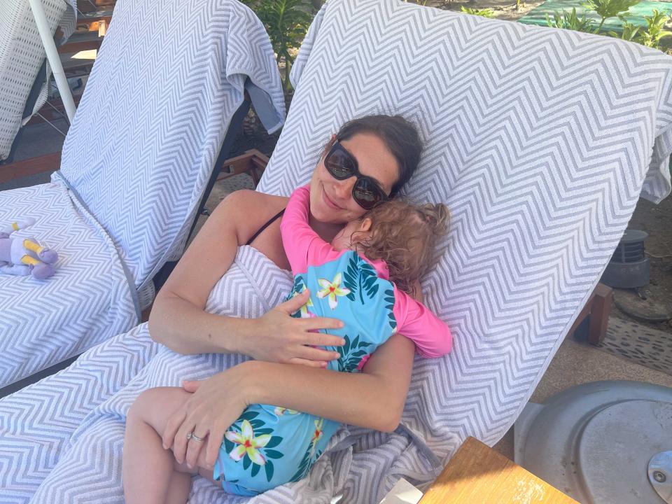 A woman wearing sunglasses hugs her child on a pool chair covered in towels.