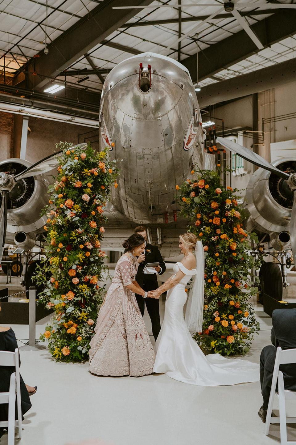 Two brides laugh at their wedding altar in front of a plane.