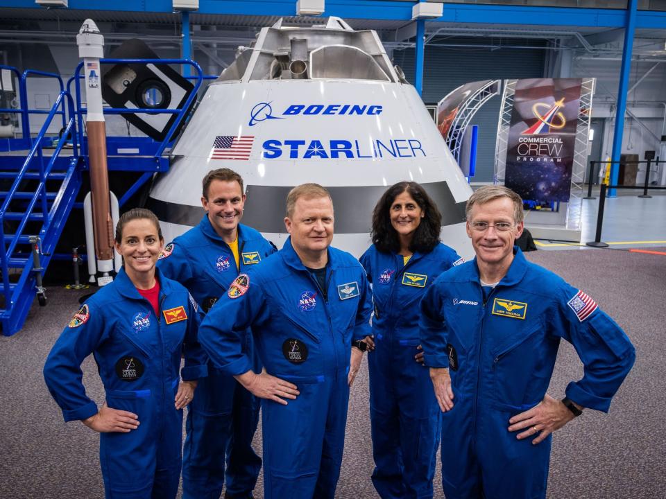 five astronauts in blue jumpsuits pose in front of starliner spaceship prototype