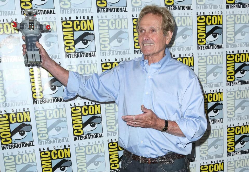 Mark Goddard holds up a robot in his right hand as he stands in front of a Comic-Con backdrop.