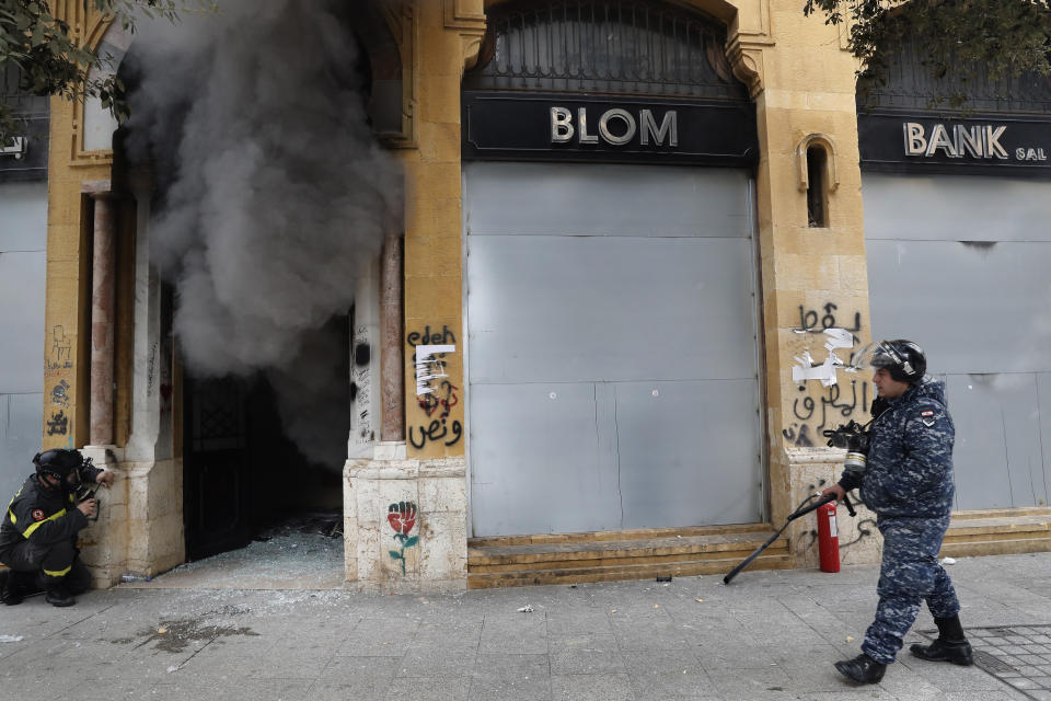 FILE - In this Tuesday, Feb. 11, 2020 file photo, a firefighter looks before entering a bank that was set on fire by anti-government protesters, as a riot policeman passes by, during a protest in downtown Beirut, Lebanon. Fitch Ratings said Tuesday, Feb. 18, 2020, that Lebanon's financial position points to a likely restructuring of the country's massive debt and financial sector as the Arab nation passes though its worst financial and economic crisis in decades. (AP Photo/Hussein Malla, File)