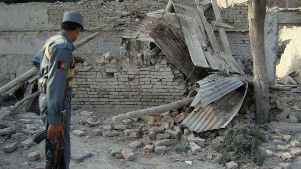 An Afghan police officer holding a semiautomatic rifle looks at a severely damaged brick wall and bent corrugated metal roof, among other rubble.