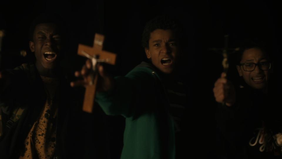 a trio of young boys stand against a black background and scream as they hold up crucifixes