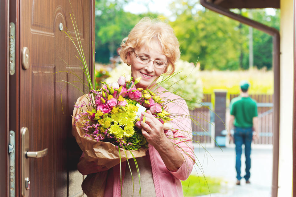 Happy senior woman standing in an entance door, holding a bouquet of flowers and reading label. Delivery man in the background. 