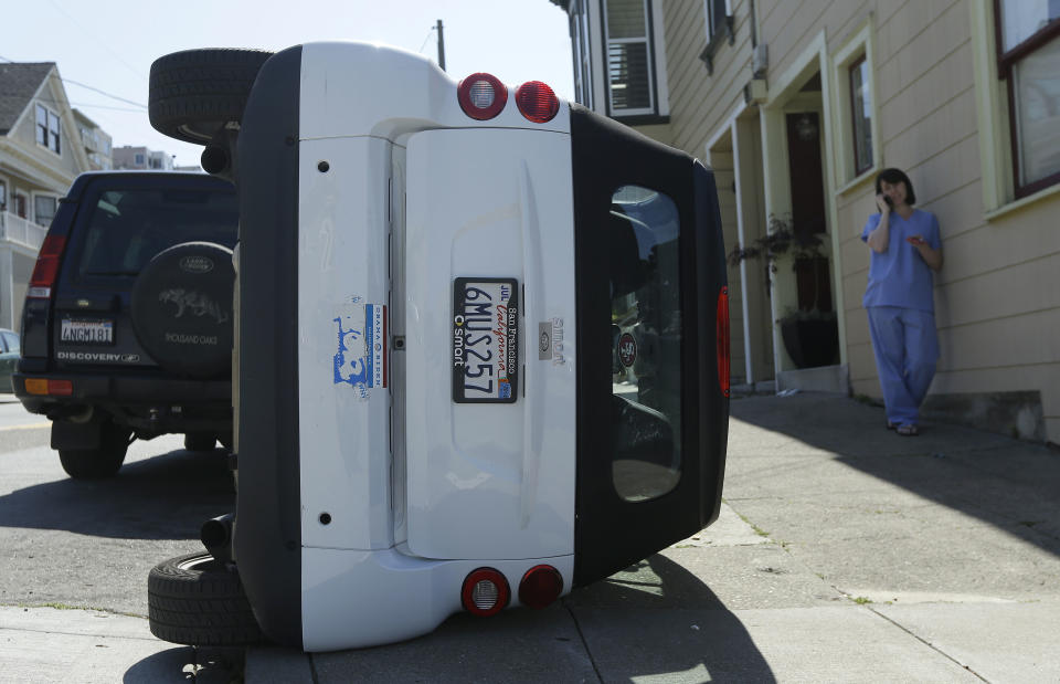 Shelley Gallivan, right, talks on the phone next to a tipped over Smart car which belongs to her friend on the corner of Prospect and Oso streets in San Francisco, Monday, April 7, 2014. Police in San Francisco are investigating why four Smart cars were flipped over during an apparent early morning vandalism spree. Officer Gordon Shyy, a police spokesman, says the first car was found flipped on its roof and a second was spotted on its side around 1 a.m. Monday in the Bernal Heights neighborhood. (AP Photo/Jeff Chiu)