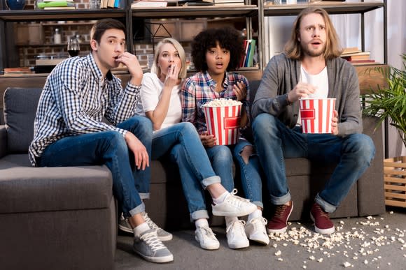 Four young people with shocked expressions are eating popcorn and watching a movie.