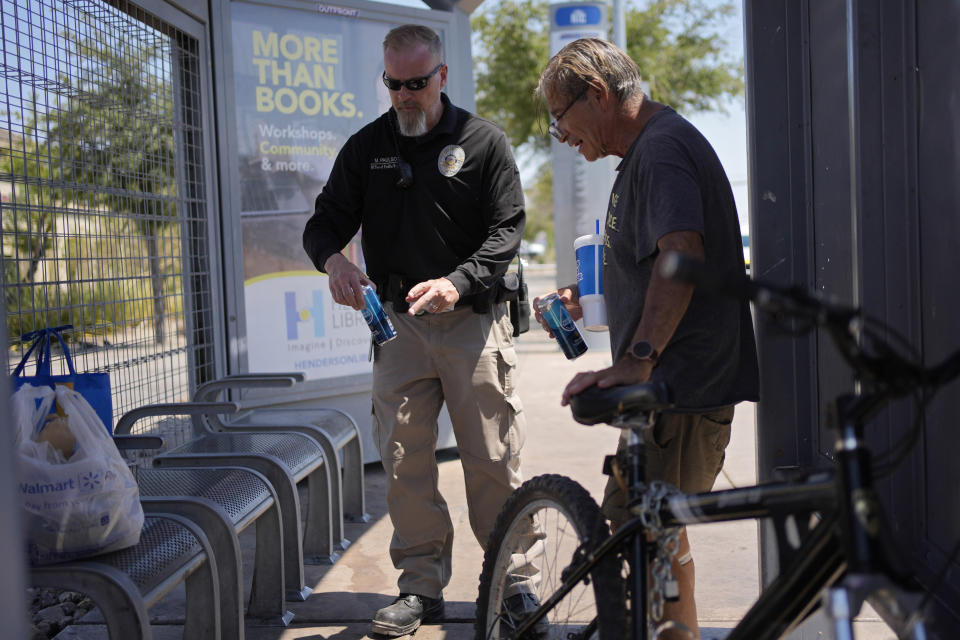 Mark Paulson, a Public Response and Code Enforcement officer, gives cans of cold water to Russell Eibeck, Wednesday, July 10, 2024, in Henderson, Nev. About 14 officers from the Office of Public Response drove around the city Wednesday, offering water, electrolytes, free bus tickets, and rides to cooling centers during a heat emergency. (AP Photo/John Locher)