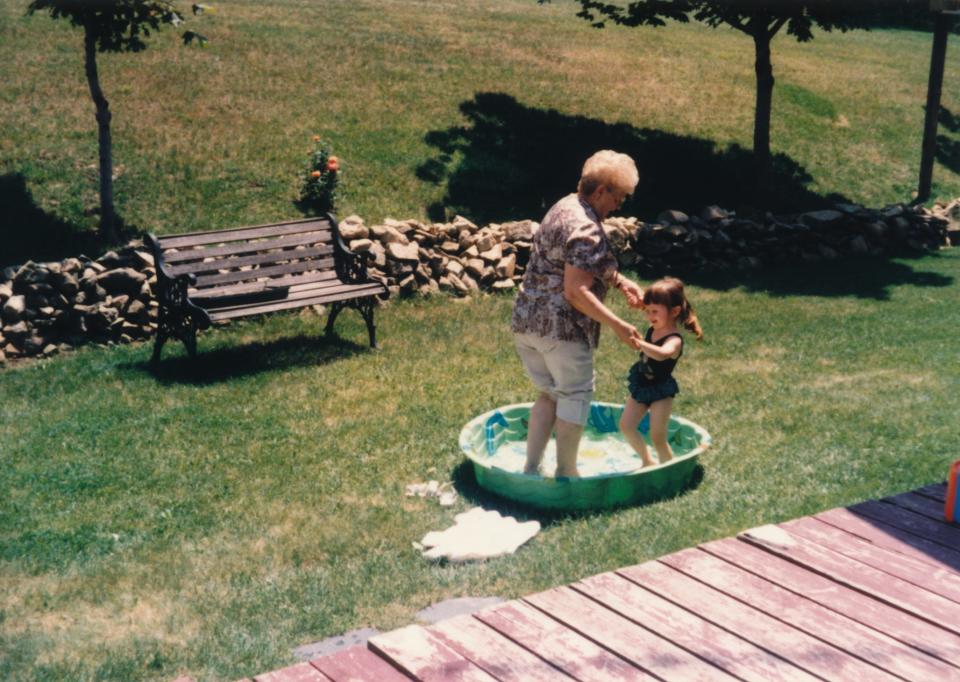 An elderly woman and a young girl play in a small green kiddie pool in a grassy backyard. A bench and a stone wall are in the background