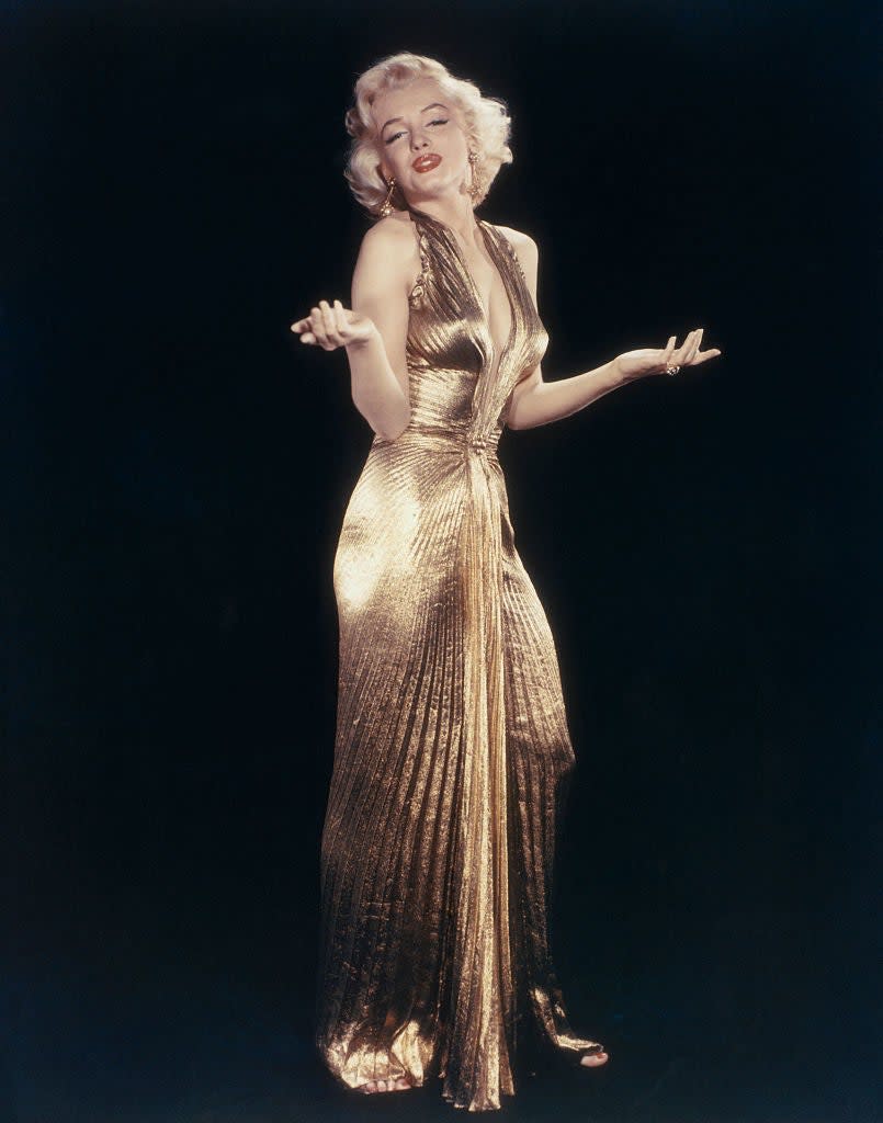 Marilyn Monroe poses with a playful expression, wearing a floor-length, metallic, pleated gown