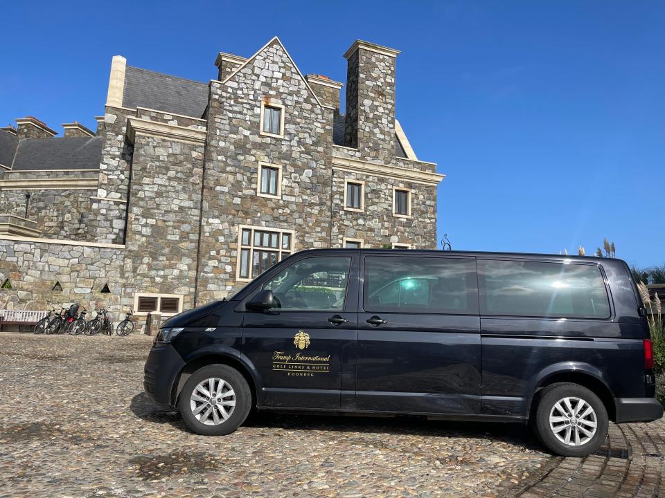 A black van with a Trump International Gold Links and Hotel design on the side in front of hotel with gray stone