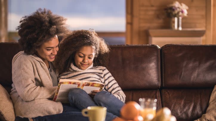 happy black mother and daughter relaxing on sofa at home and reading a book