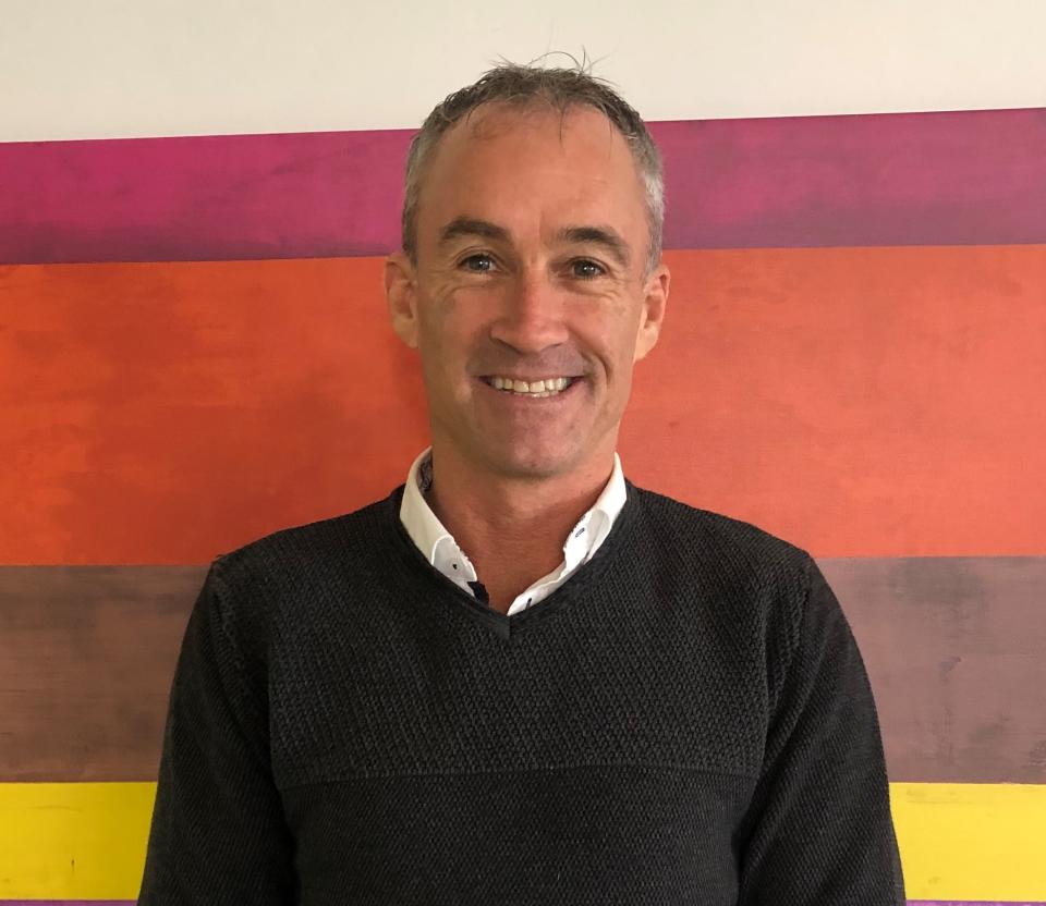 A portrait photo of Noel Stowers smiling in front of a colourful wall. 