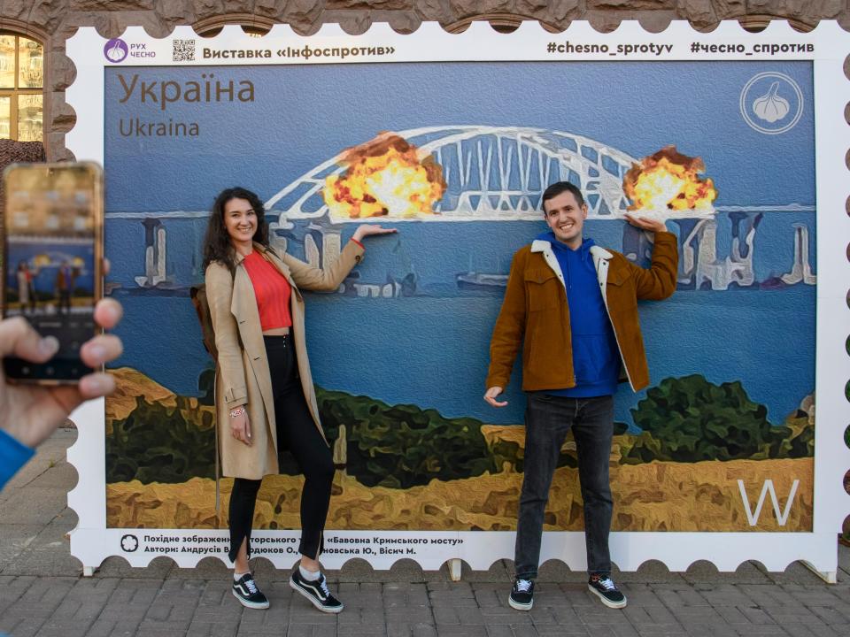 People pose for photos and take selfies in front of the large poster form of postage stamp depicting the Crimean Kerch Bridge on fire in Kyiv, Ukraine, October 08, 2022.