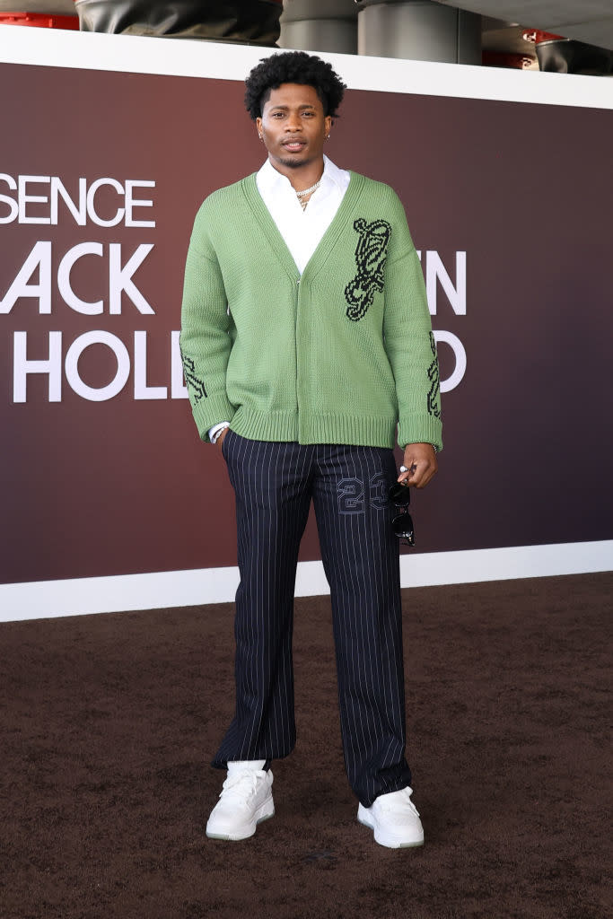 Da'Vinchi stands on the red carpet, wearing a cardigan over a white, striped pants, and sneakers