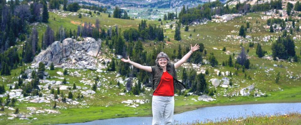 Woman spreads her arms wide to embrace the scenic beauty of Beartooth Pass in Wyoming.  She has on a red shirt and khaki pants.