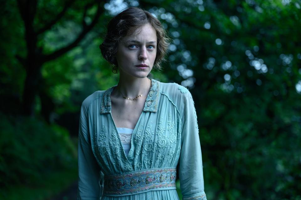 Emma Corrin in a vintage-style dress with intricate embroidery, standing outdoors with trees in the background