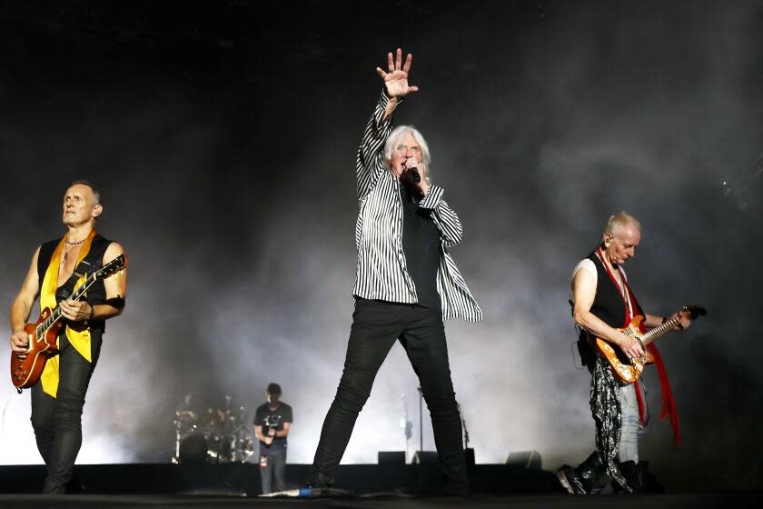 Three men on a stage -- two men playing guitars on either side of a man singing into a microphone and reaching his hand out