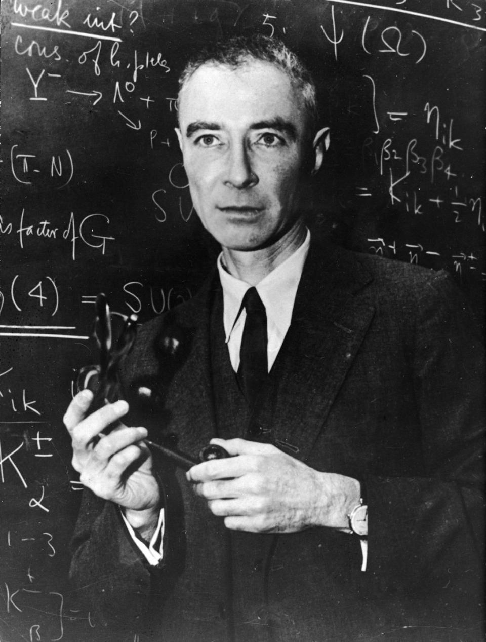 j robert oppenheimer wears a suit and tie and stands in front of a chalkboard with scientific problems written on it