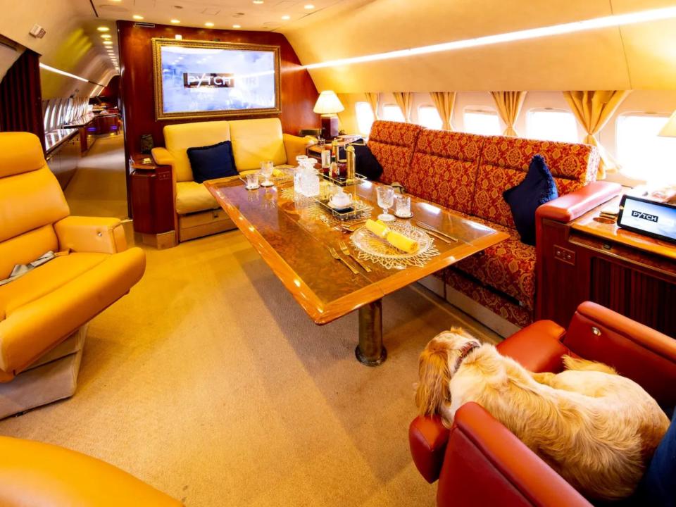 The main lounge with TV on, red couch, wood dining table, and beige and red armchairs.