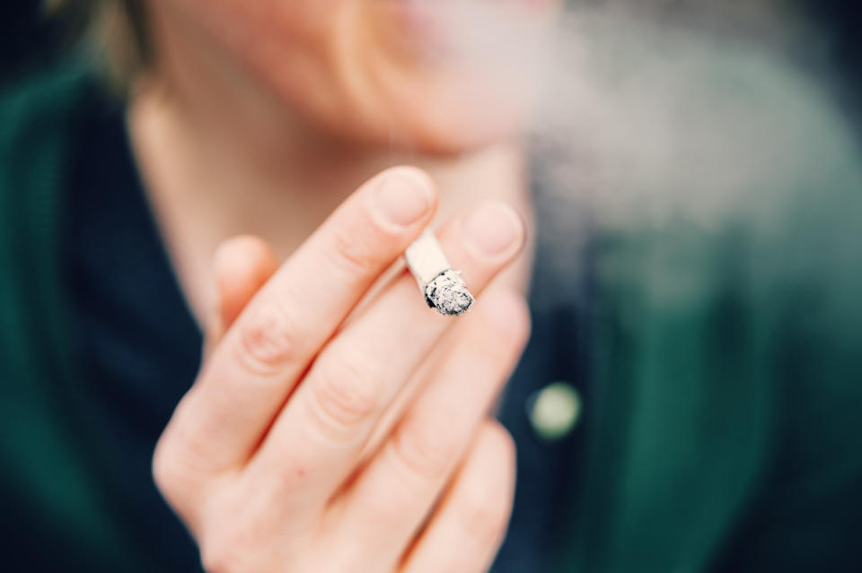 A person holding a lit cigarette close to their mouth, with smoke visible. The image focuses on the hand and cigarette