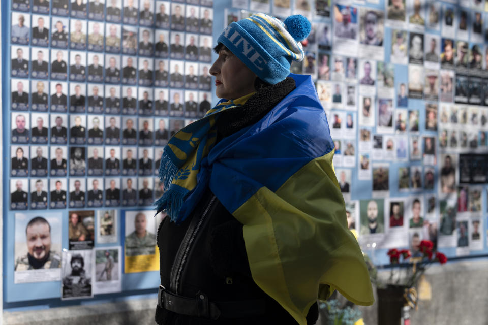 A woman wrapped in a Ukrainian flag stands next to the Memory Wall of Fallen Defenders of Ukraine in Russian-Ukrainian War on Ukrainian Volunteer Day in Kyiv, Ukraine, Tuesday, March 14, 2023. (AP Photo/Andrew Kravchenko)