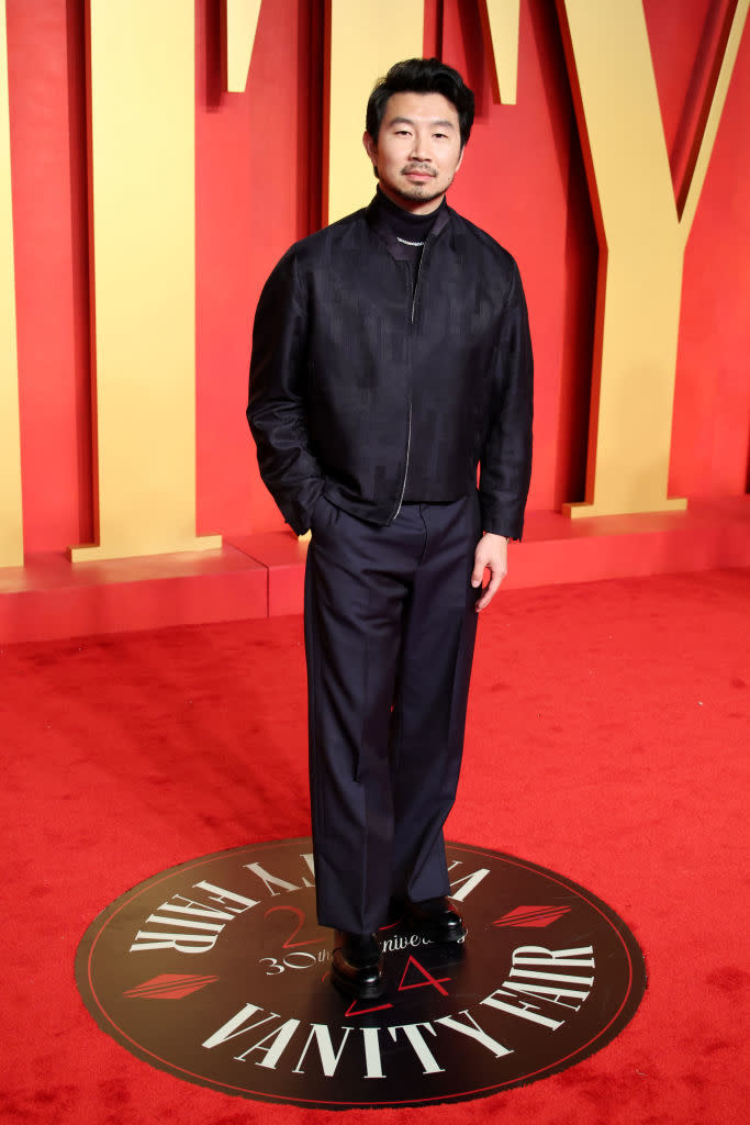 Man on red carpet in black jacket, turtleneck, and trousers, hands in pockets