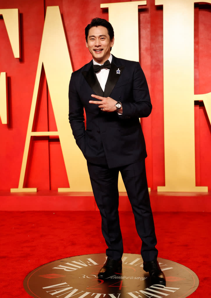 Man in tuxedo standing on red carpet, styled hair, smiling, and making a peace sign