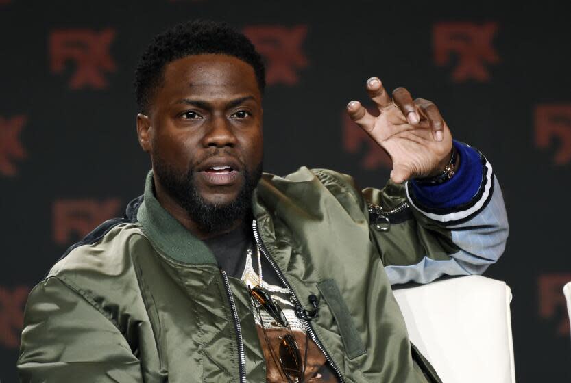 Kevin Hart in an olive-drab jacket raising his hand