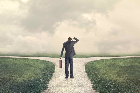 Man standing at a crossroads with a briefcase in his left hand and scratching his head under a sky full of white clouds