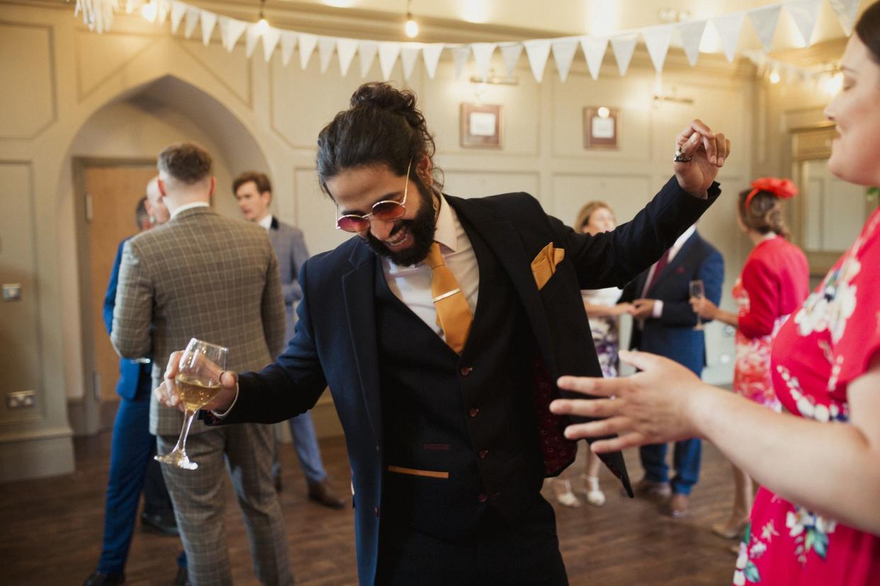 Man at a wedding dressed in a suit and sunglasses dancing while other guests stand around him. He is holding a glass of wine and laughing.