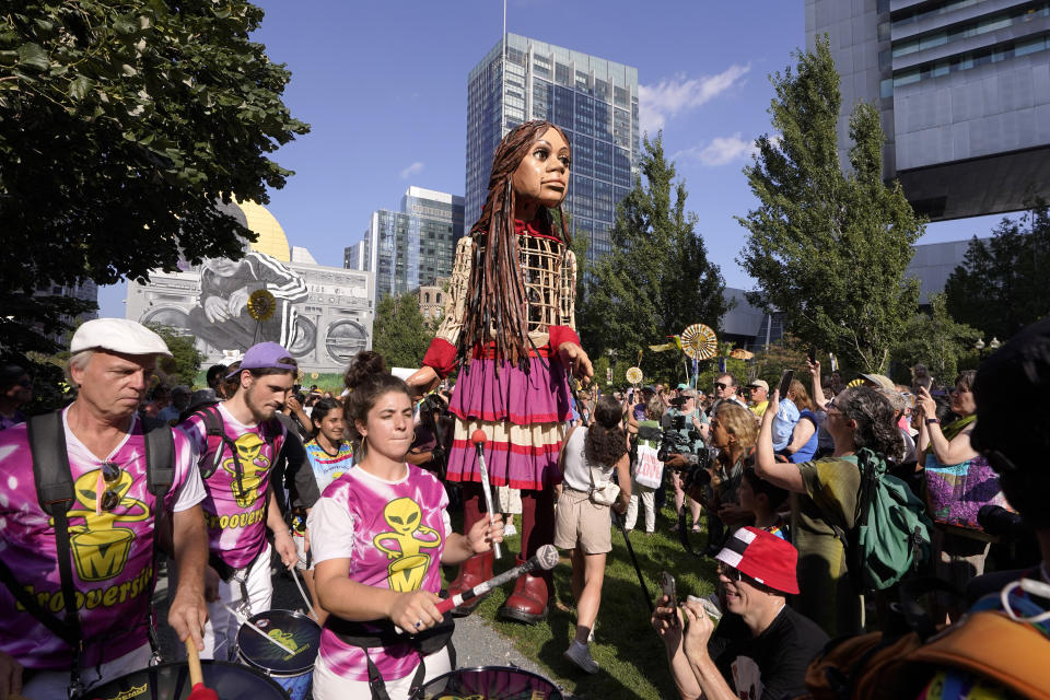 Little Amal, a 12-foot puppet of a 10-year-old Syrian refugee girl, center, is greeted by a crowd, including musicians, left, while walking with the assistance of puppeteers, Thursday, Sept. 7, 2023, in Boston. The puppet is scheduled to journey across the United States, with planned stops in over 35 towns and cities, between Sept. 7 and Nov. 5, 2023, in an effort to raise awareness about refugees and displaced people across the world. (AP Photo/Steven Senne)