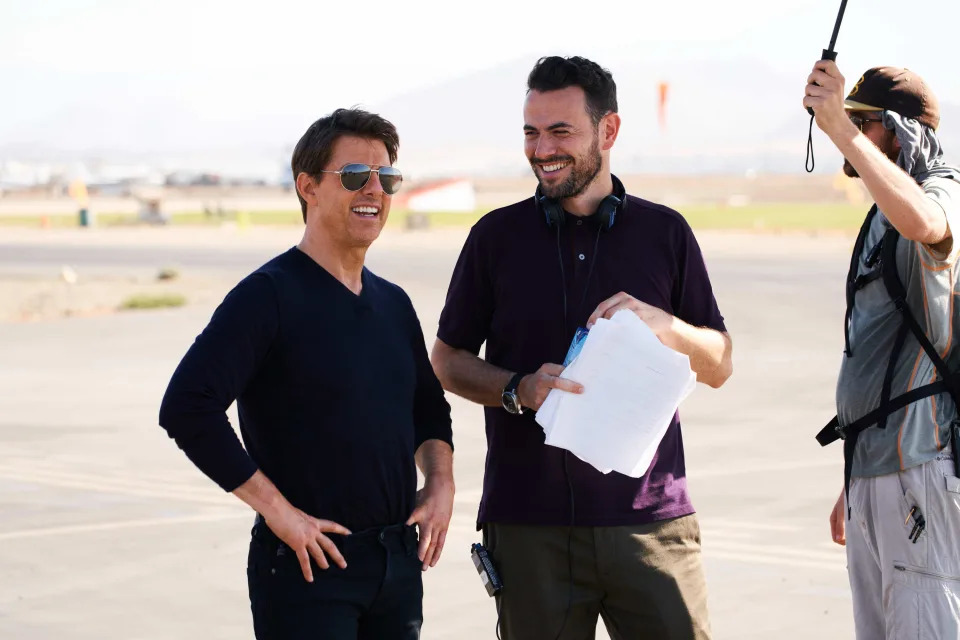 Tom Cruise, in casual wear, chats with a crew member holding a script on an outdoor set. Another crew member adjusts equipment