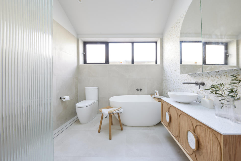 A shot of the bathroom with a toilet on the left, a bathroom on the right and a wooden vanity on the right wall with a mirror above it. 