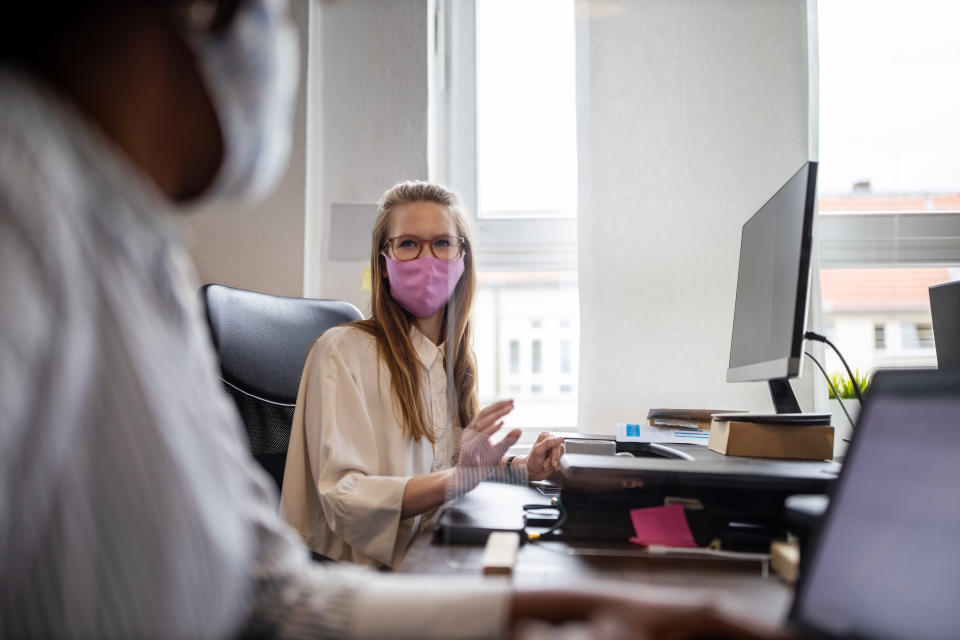 Businesswoman discussing work through glass partition with colleague at office desk. Business people back to work sitting at desk with protection guard between them post covid-19 pandemic.