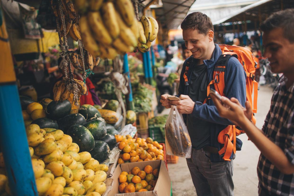 Photo of a tourist, on a local flea market, using a mobile phone while buying vegetables and fruit