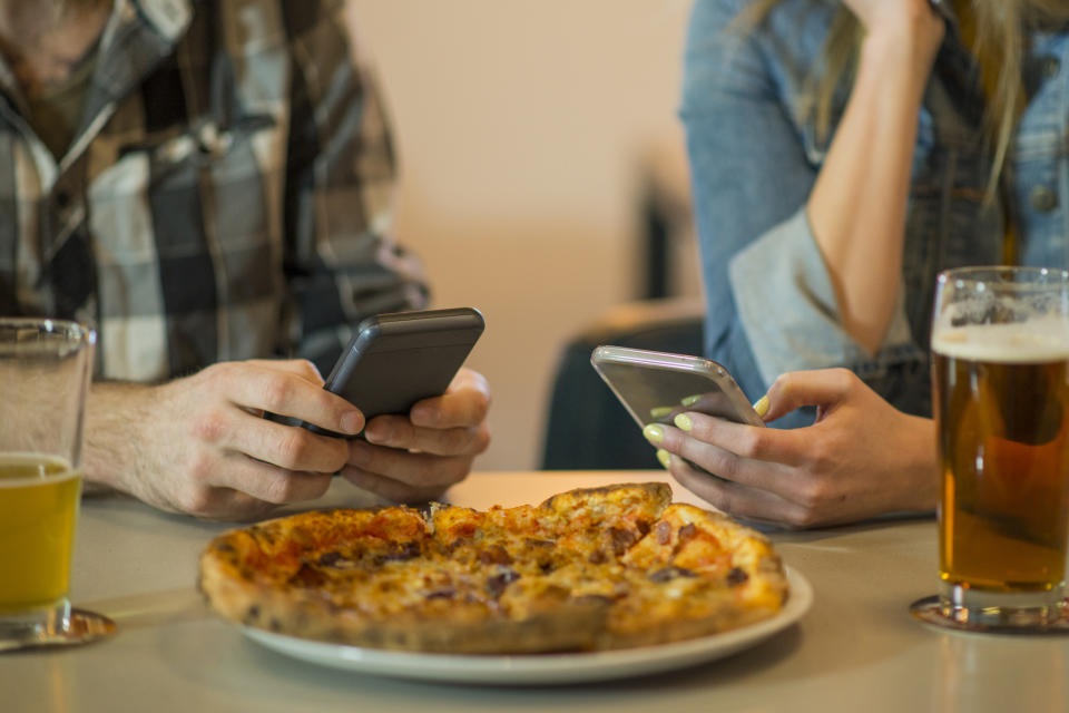 A man and a woman sitting at a table with phones, not engaging with each other, and a pizza and beer in front of them