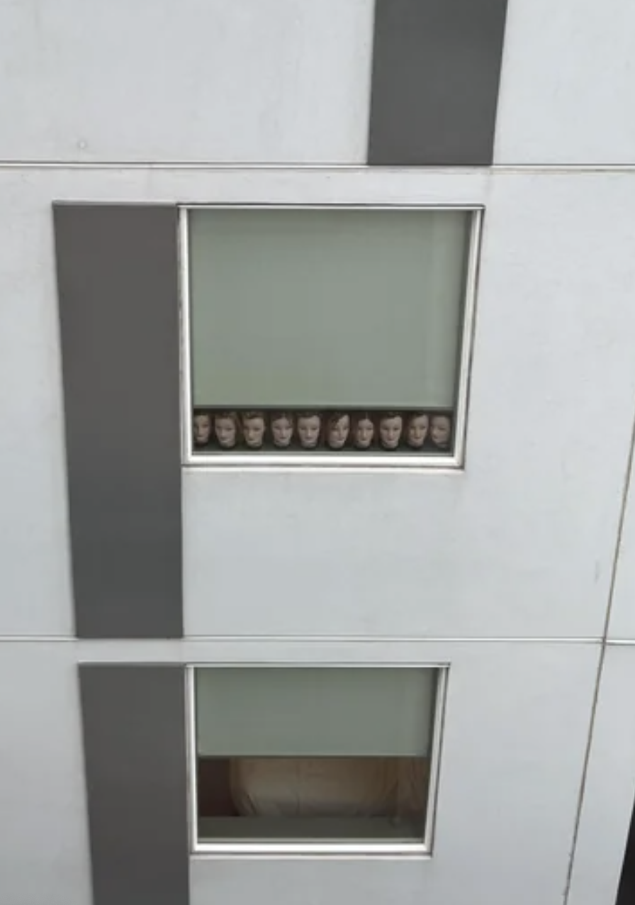 Multiple people are seen through a square window, appearing to be in a line, with a bed visible in the lower window