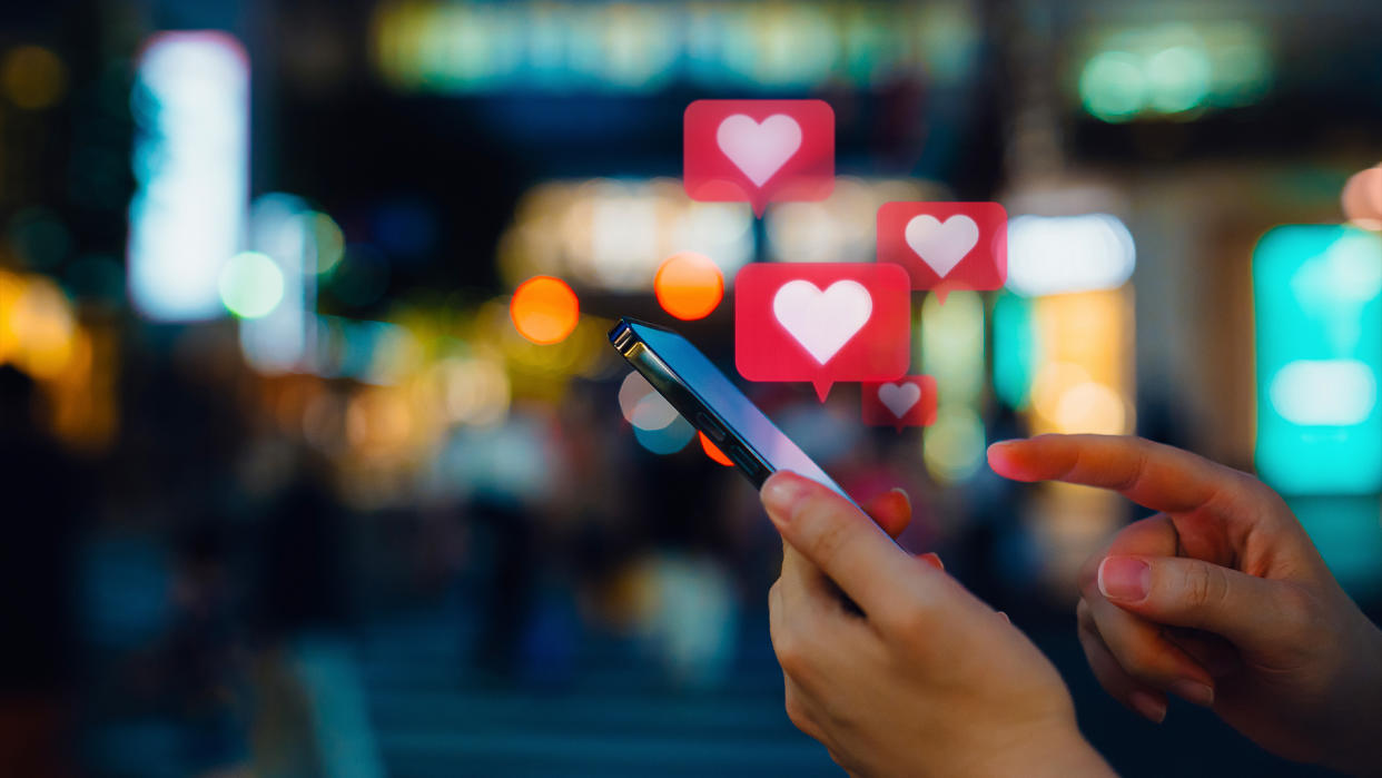  Edited photo shows a woman's hands as she lifts a finger to swipe on a phone. Hearts in speech bubbles are shown popping up above the phone, indicating likes or notifications. 