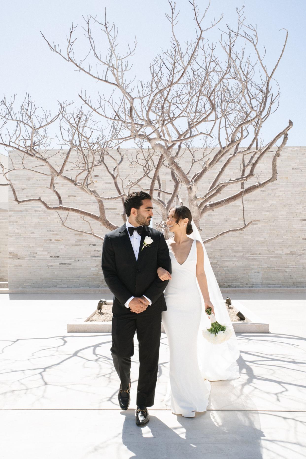 A bride and groom pose in their wedding attire with their arms locked in front of a tree.