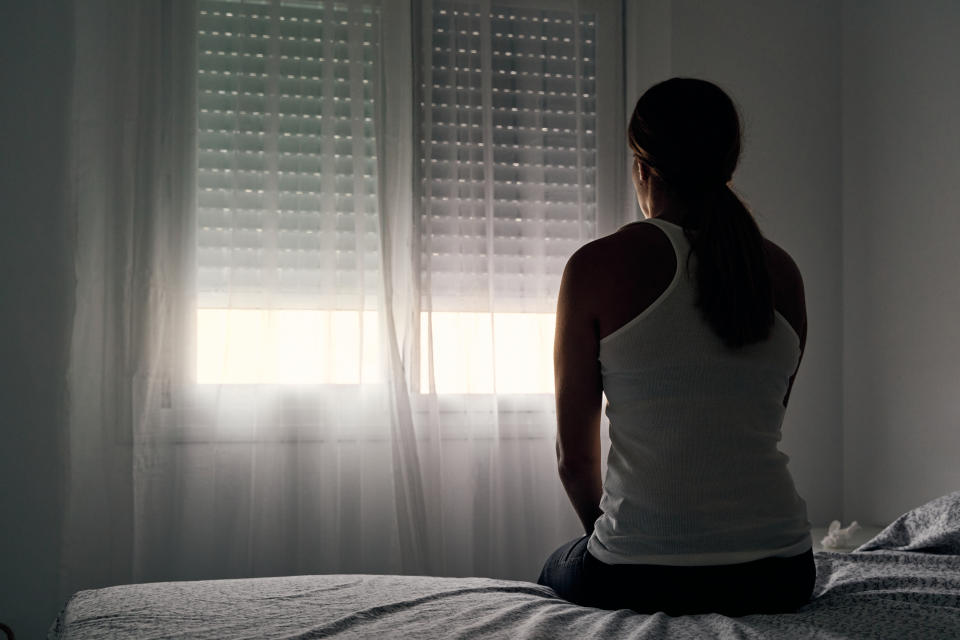 Woman sitting on a bed facing a window with closed shutters, room appears dimly lit