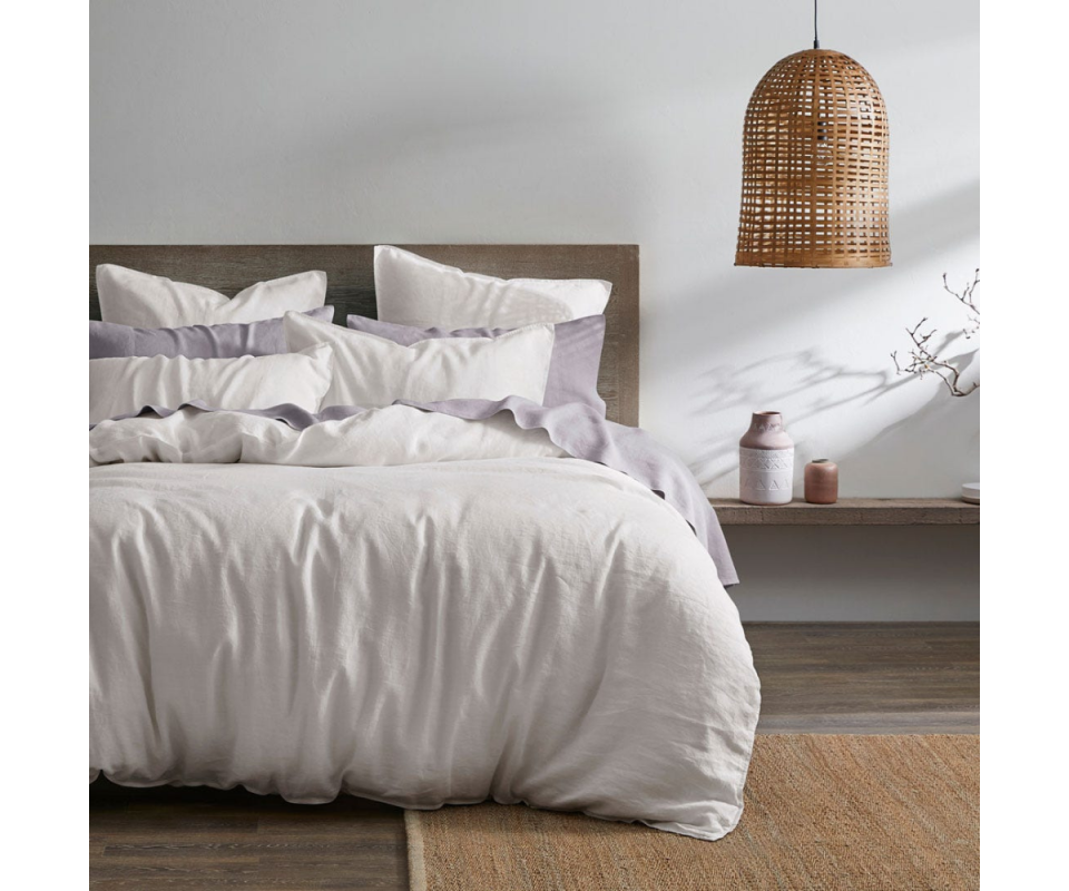 A white queen bed with a white linen quilt and a straw hanging pendant lamp on the right with wooden flooboards beneath