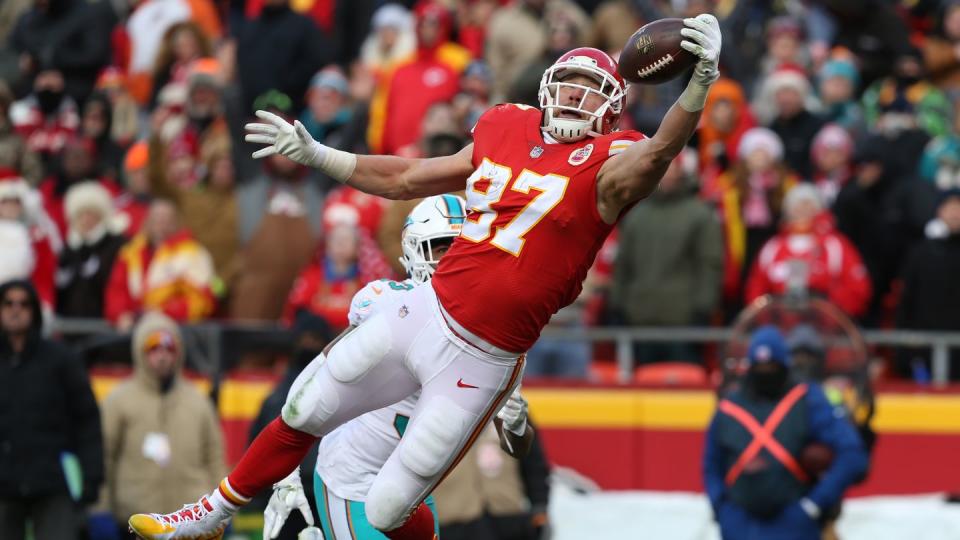 travis kelce, wearing a kansas city chiefs football uniform, jumping in the air and trying to catch a football with one hand