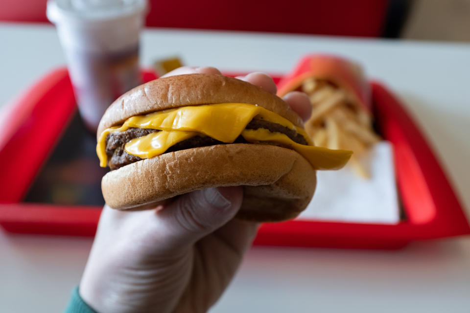 Person holding a cheeseburger over a tray with fries and a drink in the background