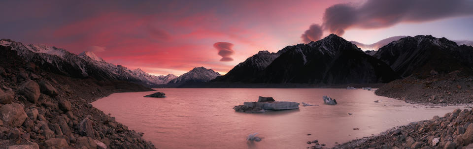 A pinky-reddish sunrise view at Tasman Glacier in Mount Cook National Park with lenticular clouds hovering all above the Mount Cook mountain range.