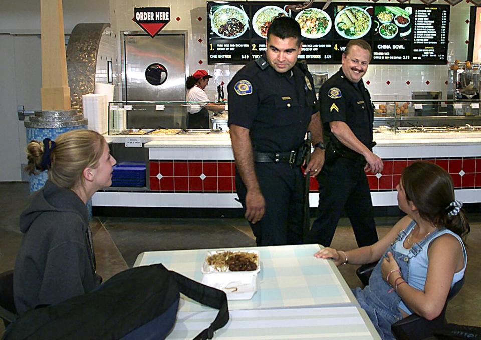 Two police officers in uniform walk by two people at a table in a food court.